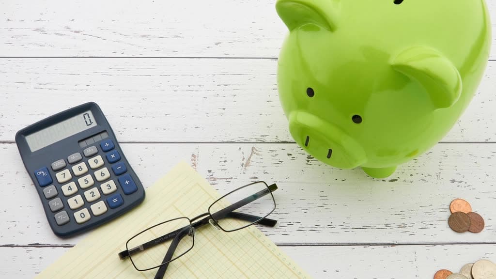 Green piggy bank beside a calculator, reading glasses and a notepad on a white wood table